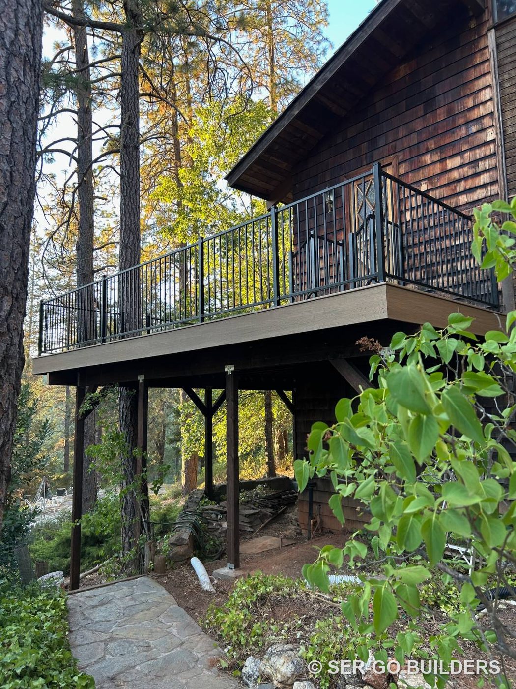 Second-story composite deck with black steel railing wrapping a cedar-shingle home among pine trees